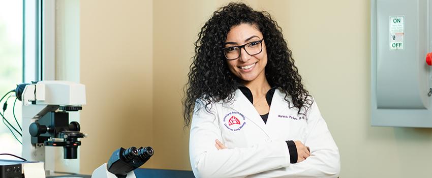 Female student in white lab coat with microscope behind her.