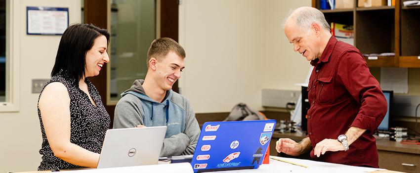 Two professors smiling with student in classroom looking at laptop