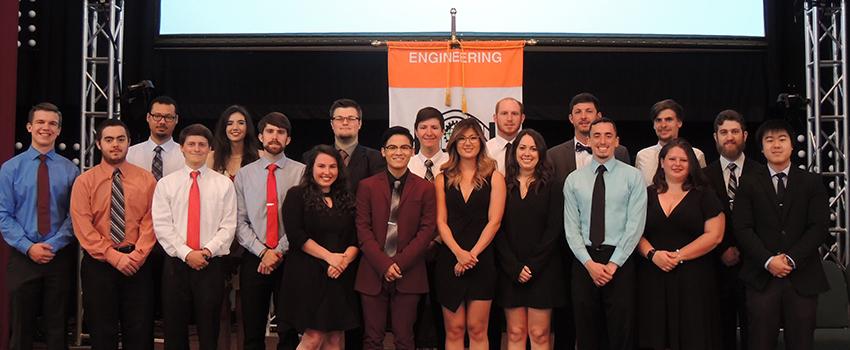 Engineering students standing on stage in front of flag.