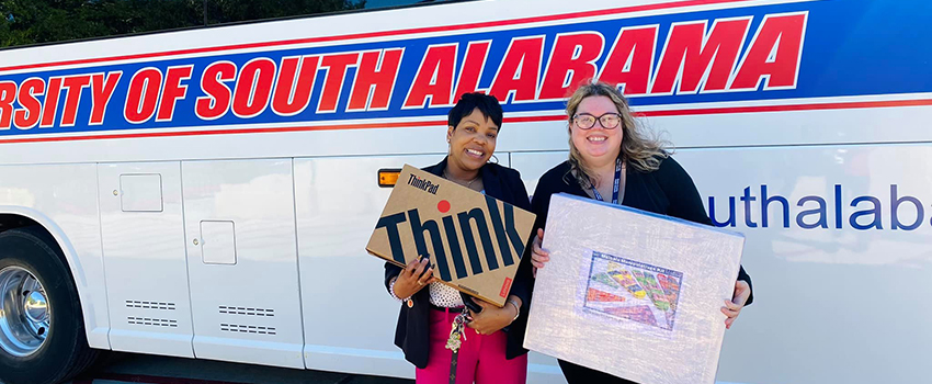 Two people smiling in front of USA bus holding classroom materials.