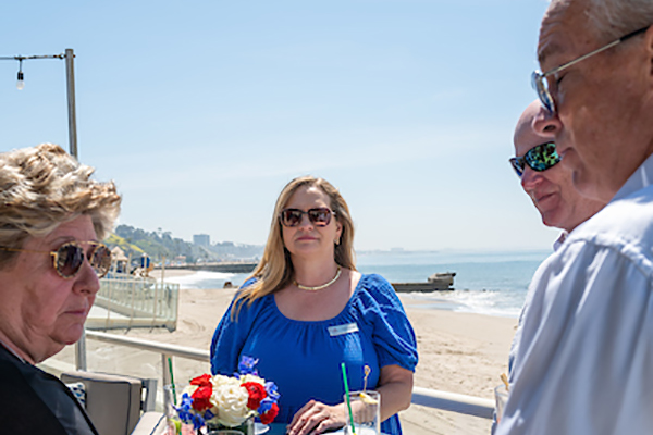 Woman in blue dress listening to other alumni with the beach in the background.