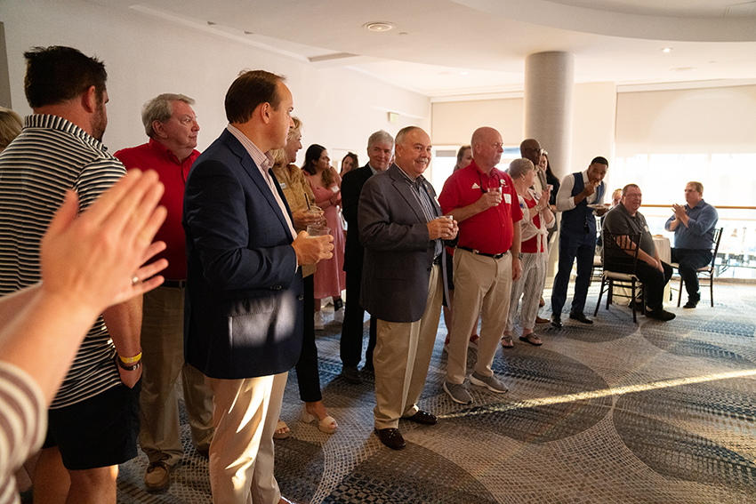 Man in red shirt speaking with group of alumni clapping.