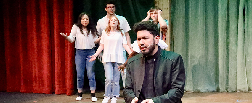 USA Students acting on stage in a play with one male student in front kneeling and 4 students behind him.