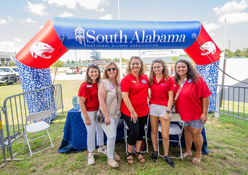 Five alumni smiling in front of a University of South Alabama sign.