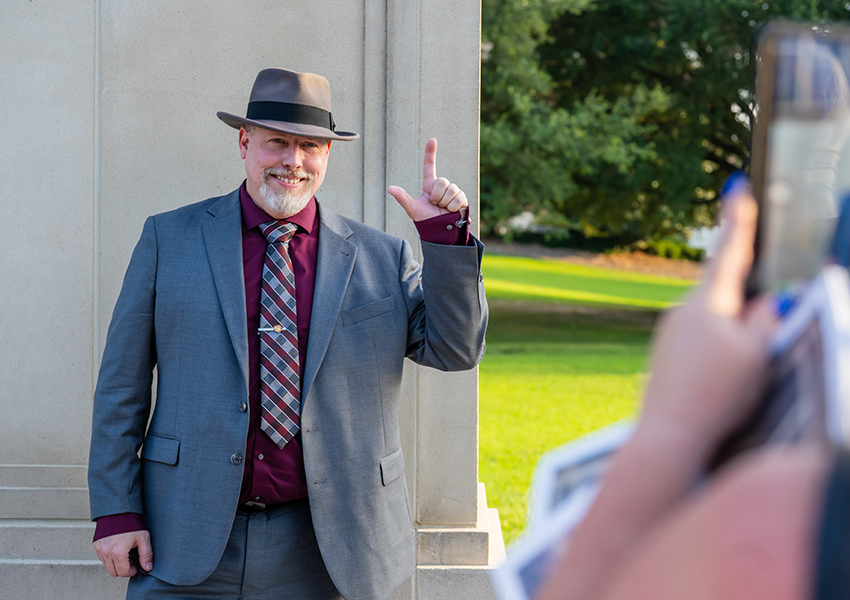 Man in hat holding up J sign in front of Wall of Honor.