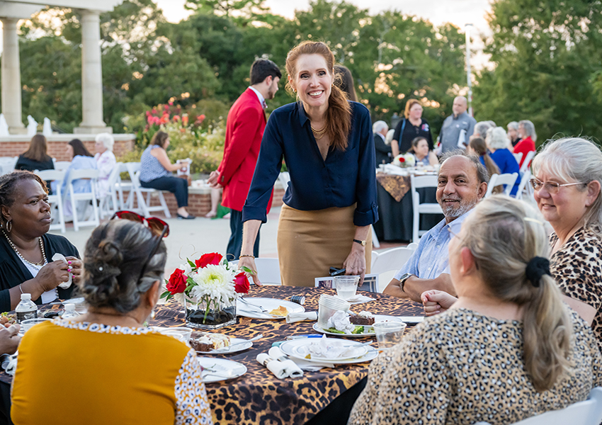 Woman standing behind table smiling speaking to people sitting at table.