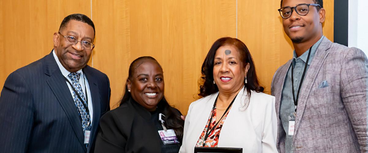 Four people stand together indoors, with one woman in the center holding an award plaque. All are smiling and dressed in business attire.