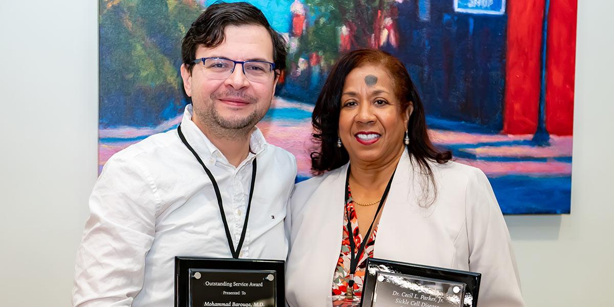 Two people standing side by side, each holding a plaque award, posing for a photo in front of a colorful painting.