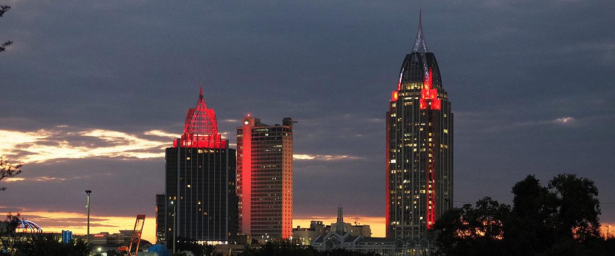Landscape of downtown Mobile around sunset on a cloudy day. Battle House and Trustmark towers with red lights.