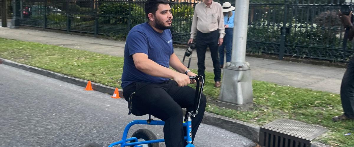 Bearded man in a blue shirt rides a blue handcycle along a city street, watched by pedestrians near a park fence and traffic cones.