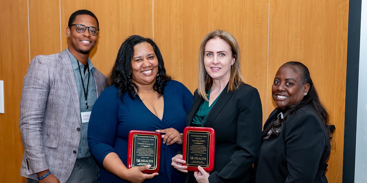 Four people stand in front of a wooden wall, two women in the center holding award plaques and smiling, while a man and another woman stand on either side of them, also smiling, at what appears to be an award ceremony.