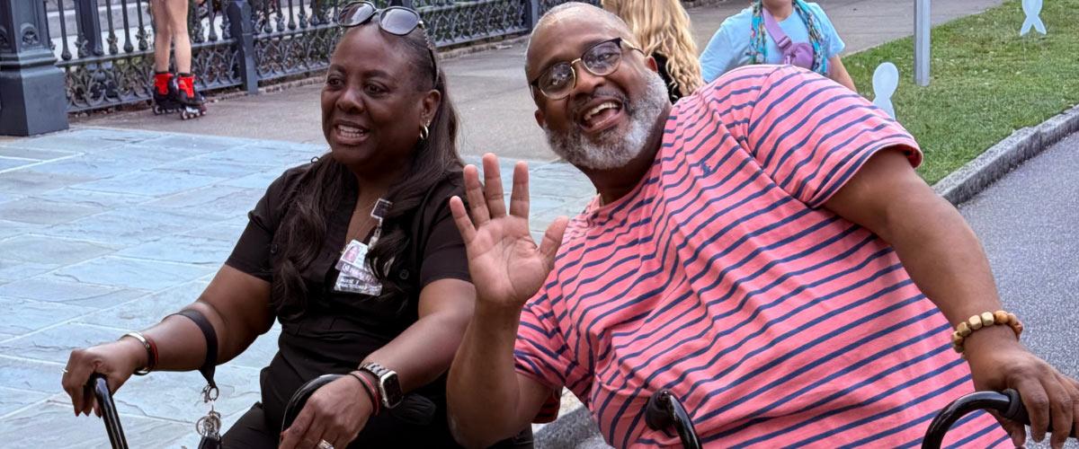 Two adults sit on mobility scooters outdoors; the man in a pink striped shirt waves and smiles at the camera, while the woman beside him also smiles. Other people and a fence are in the background.
