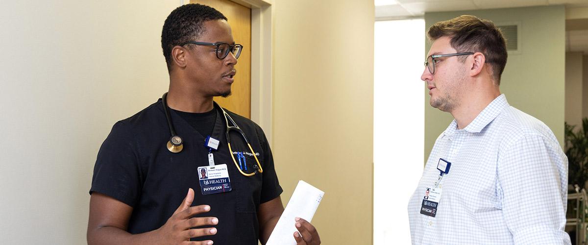 Health Physician talking with other Physician in a hallway. Physician on the left has a paper in his hand. Physician on right is listening intently.