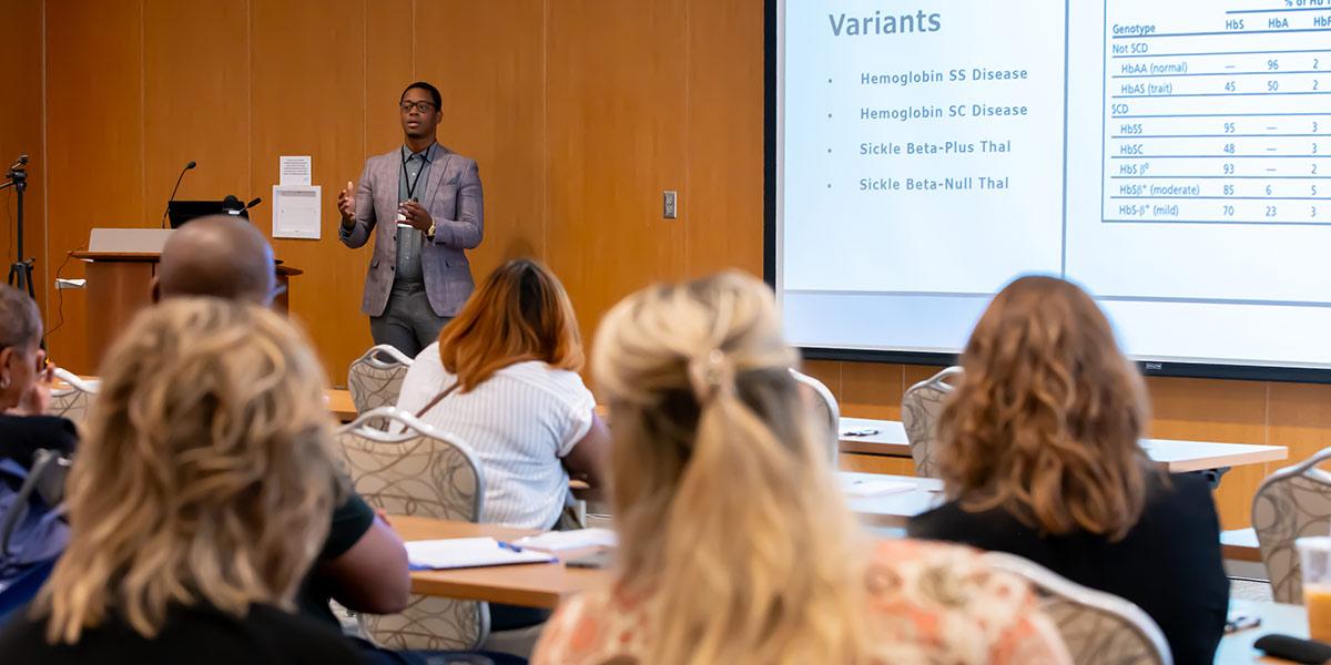 A speaker presents to an audience in a conference room. Attendees sit facing a screen displaying information about hemoglobin variants. The setting appears educational and professional.