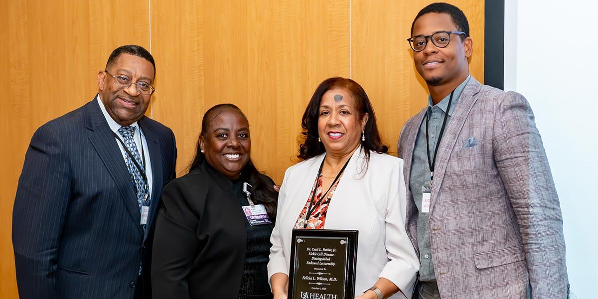 Four people stand together indoors, with one woman in the center holding an award plaque. All are smiling and dressed in business attire.