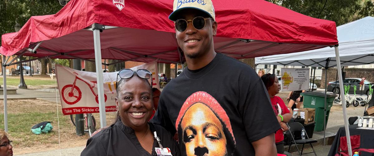 Two people stand smiling in front of red canopies at an outdoor event. The woman wears scrubs, and the man wears sunglasses and a cap. There are banners, tables, and people in the background.