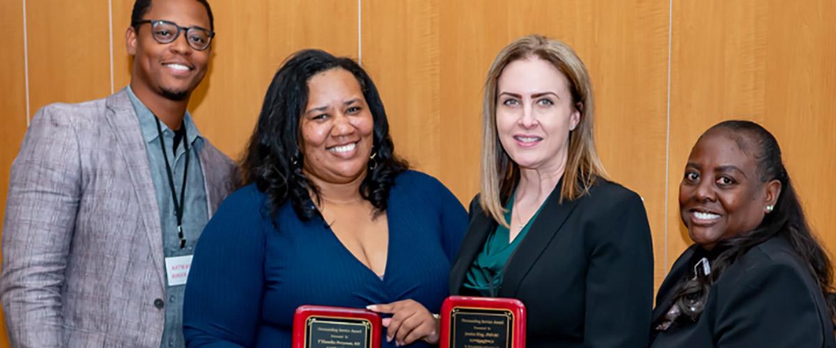 Four people stand in front of a wooden wall, two women in the center holding award plaques and smiling, while a man and another woman stand on either side of them, also smiling, at what appears to be an award ceremony.