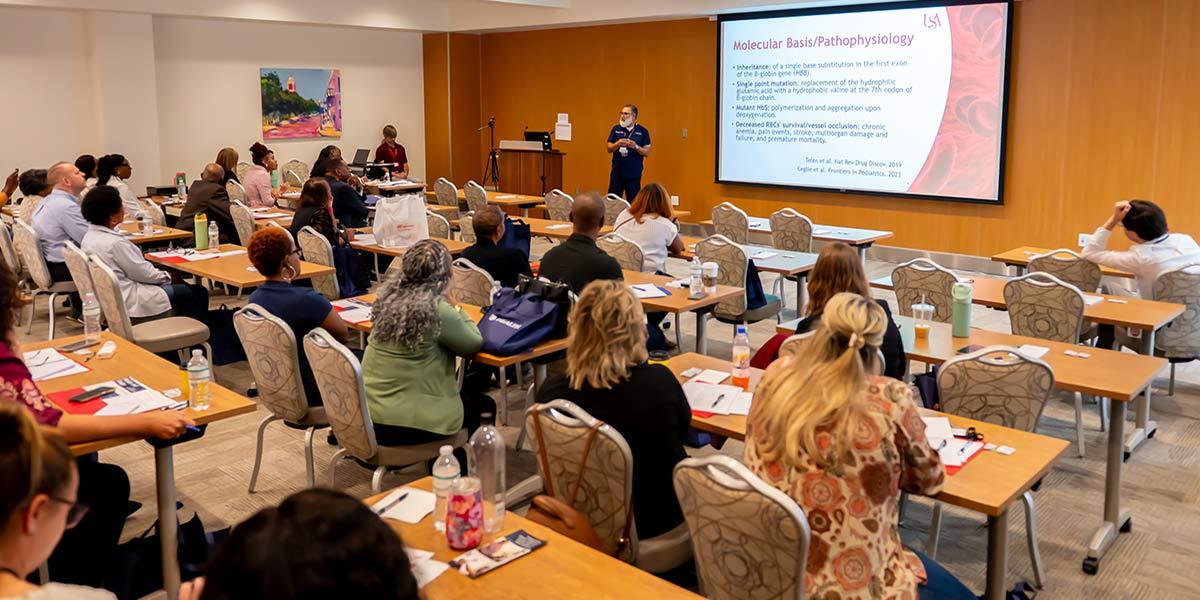 A classroom with adults seated at tables facing a presenter who is giving a lecture with a “Molecular Basis/Pathophysiology” slide projected onto a large screen at the front of the room.
