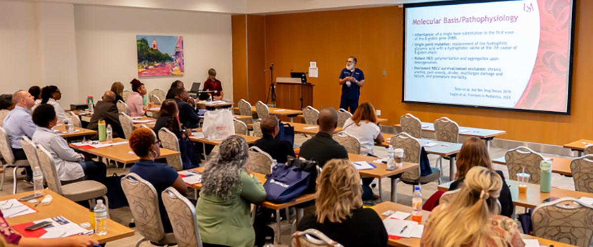 A classroom with adults seated at tables facing a presenter who is giving a lecture with a “Molecular Basis/Pathophysiology” slide projected onto a large screen at the front of the room.