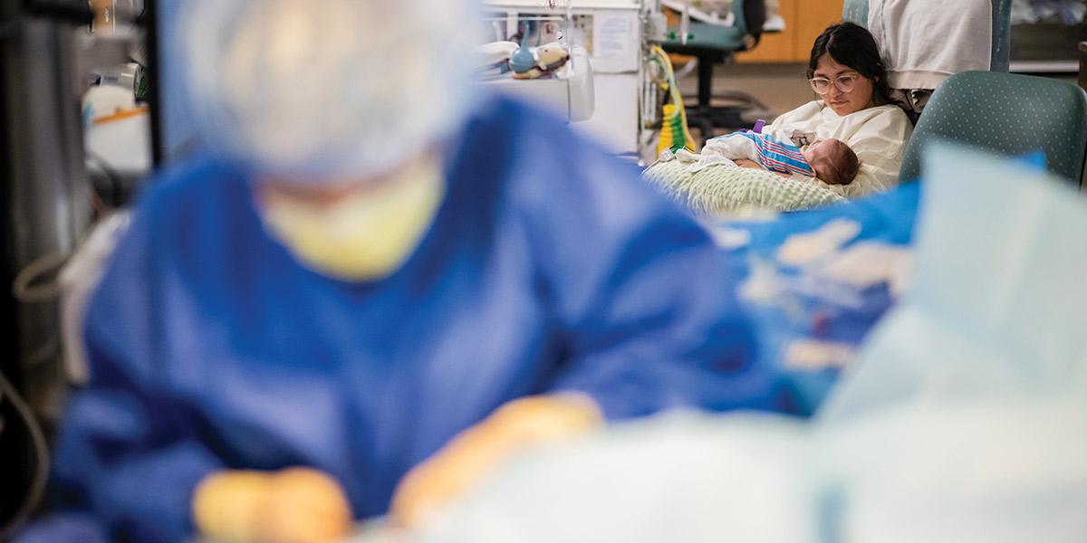 Mother holding baby in NICU with nurse working in front of her.