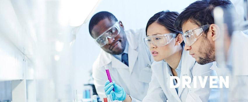 Driven text over an image of three people working in a lab with safety glasses and white coats on,