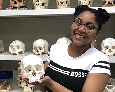 Student holding skeleton skull with rows of skulls behind her on shelves.