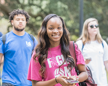 Student smiling walking outside on campus holding her cell phone.