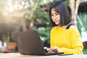 A female in glasses working on laptop outside at a table.