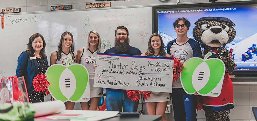 Hunter Bates receives check from Extra Yards for Teachers standing with professor, cheerleaders, and Ms. Pawla.