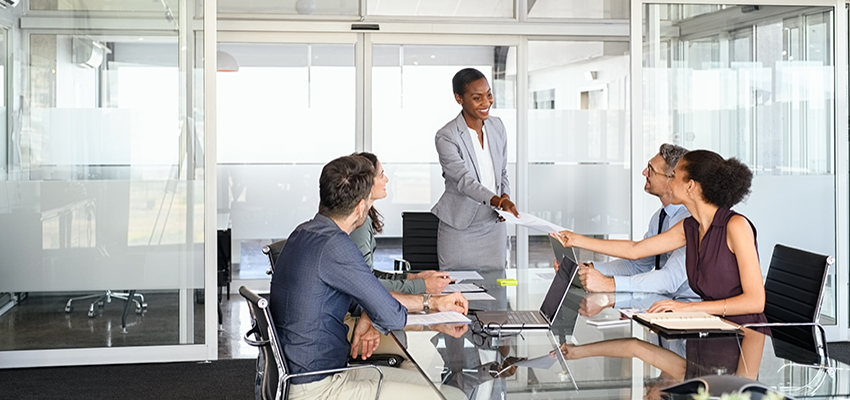 Woman smiling standing at conference table handing papers to another person sitting at the table.