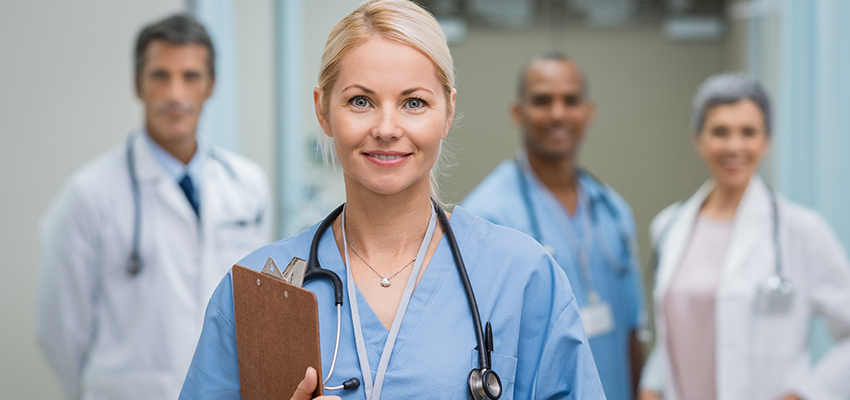 Female nurse holding clipboard with stethoscope around neck standing in front of other nurses and doctor.