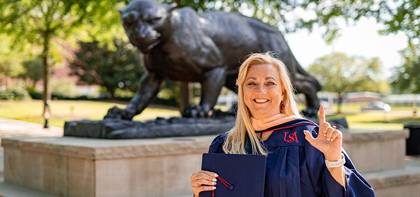 Kim Feagin standing outside on campus in cap and gown holding up the J sign for Jaguars.