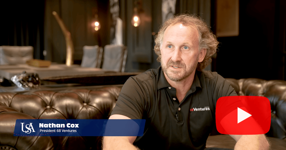 Male sits on brown leather couch during an interview. He is identified in the lower left corner as Nathan Cox