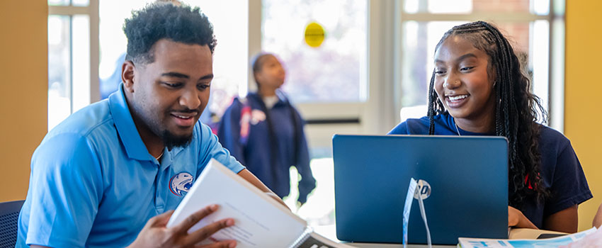 Two students looking at notebook and laptop talking.