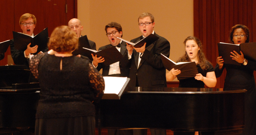 Six members of University Chorale with their director standing with music in hand as they perform on stage