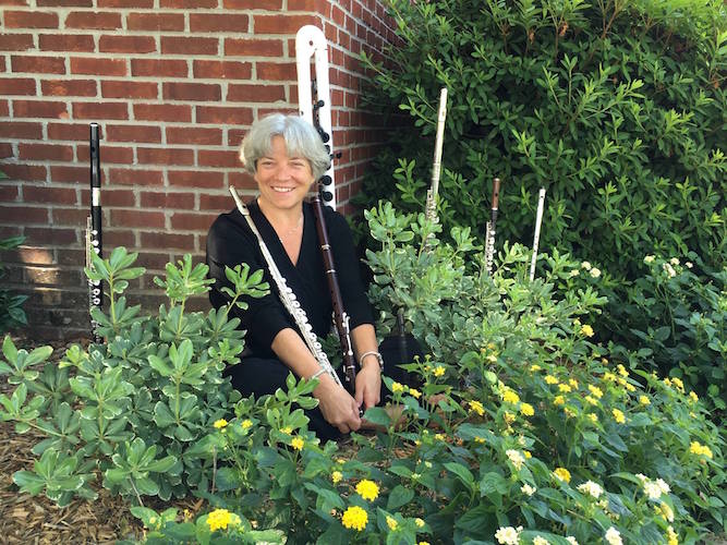 Flutist Dr. Andra Bohnet seated outdoors among yellow flowers and surrounded by several flutes