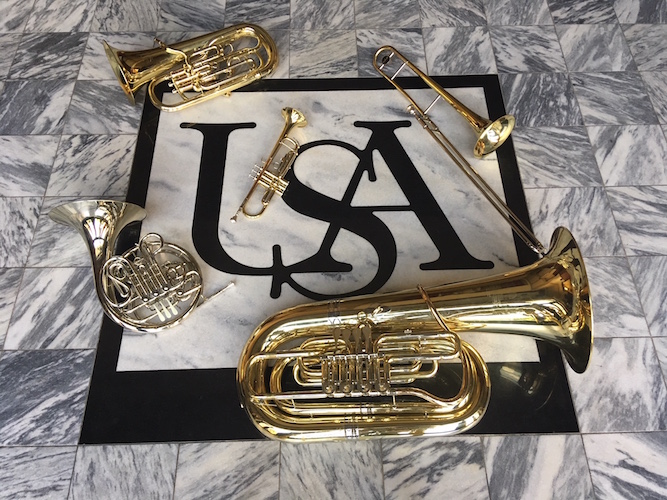 Pictured are several brass instruments laid artistically next to the USA logo on the floor of the Laidlaw Performing Arts Center.