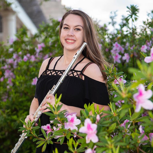 Dressed in performance attire, flutist Victoria French is pictured outdoors amongst plants on a patio. data-lightbox='featured'
