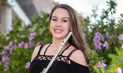 Dressed in performance attire, flutist Victoria French is pictured outdoors amongst plants on a patio.