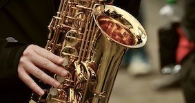 Picture is the bell of a saxophone with performer hands and fingers showing in the picture as well.