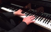 Pictured is a side-view of a grand piano keyboard with four hands performing.