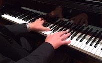 Pictured in black & white are four hands playing a piano keyboard.