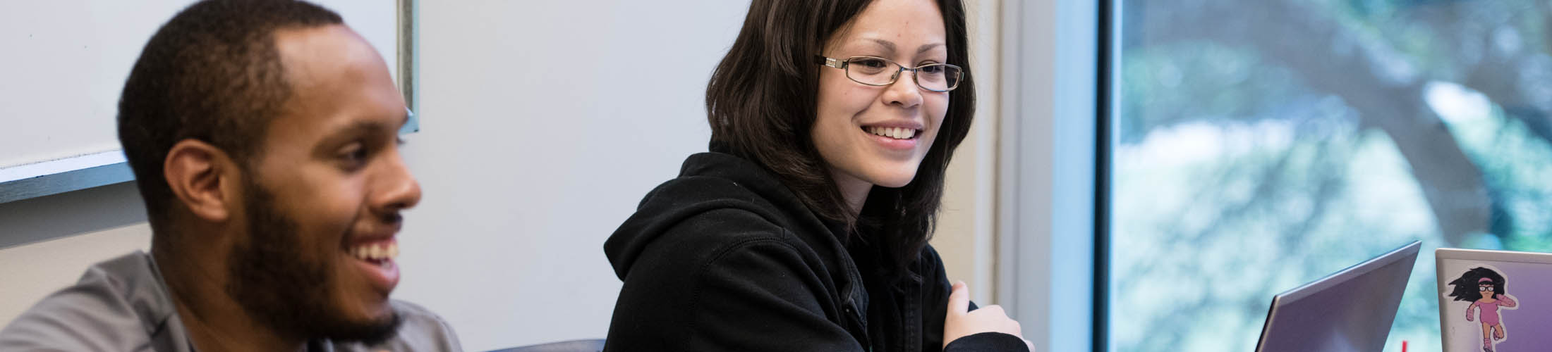 A male and female student smiling looking at their laptops in class.