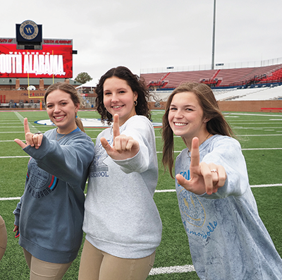 Three students holding up J sign for Jaguars on football field.