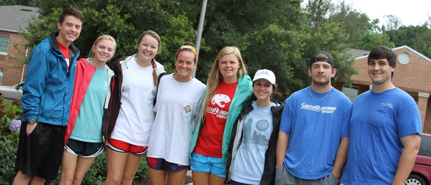 A group of students standing in front of the dorms with their arms around each other smiling.