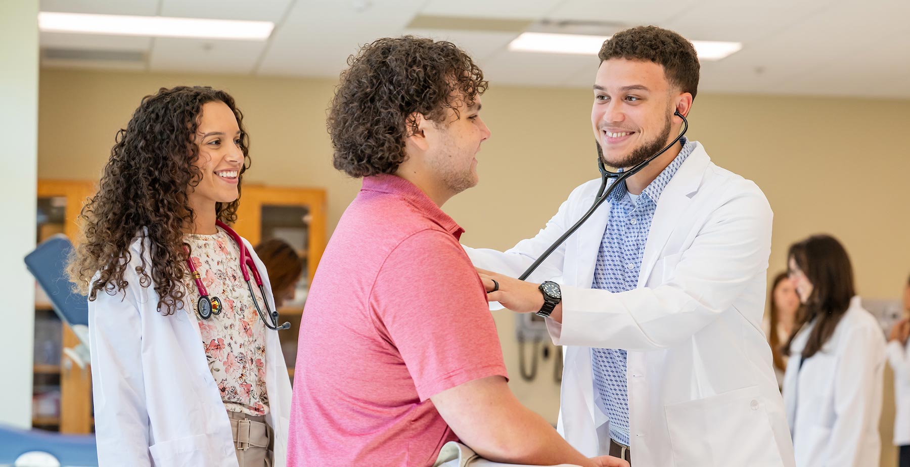 Two South Alabama Physician Assistant Studies students in white coats practice using a stethoscope with another student during a clinical skills exercise. data-lightbox='featured'