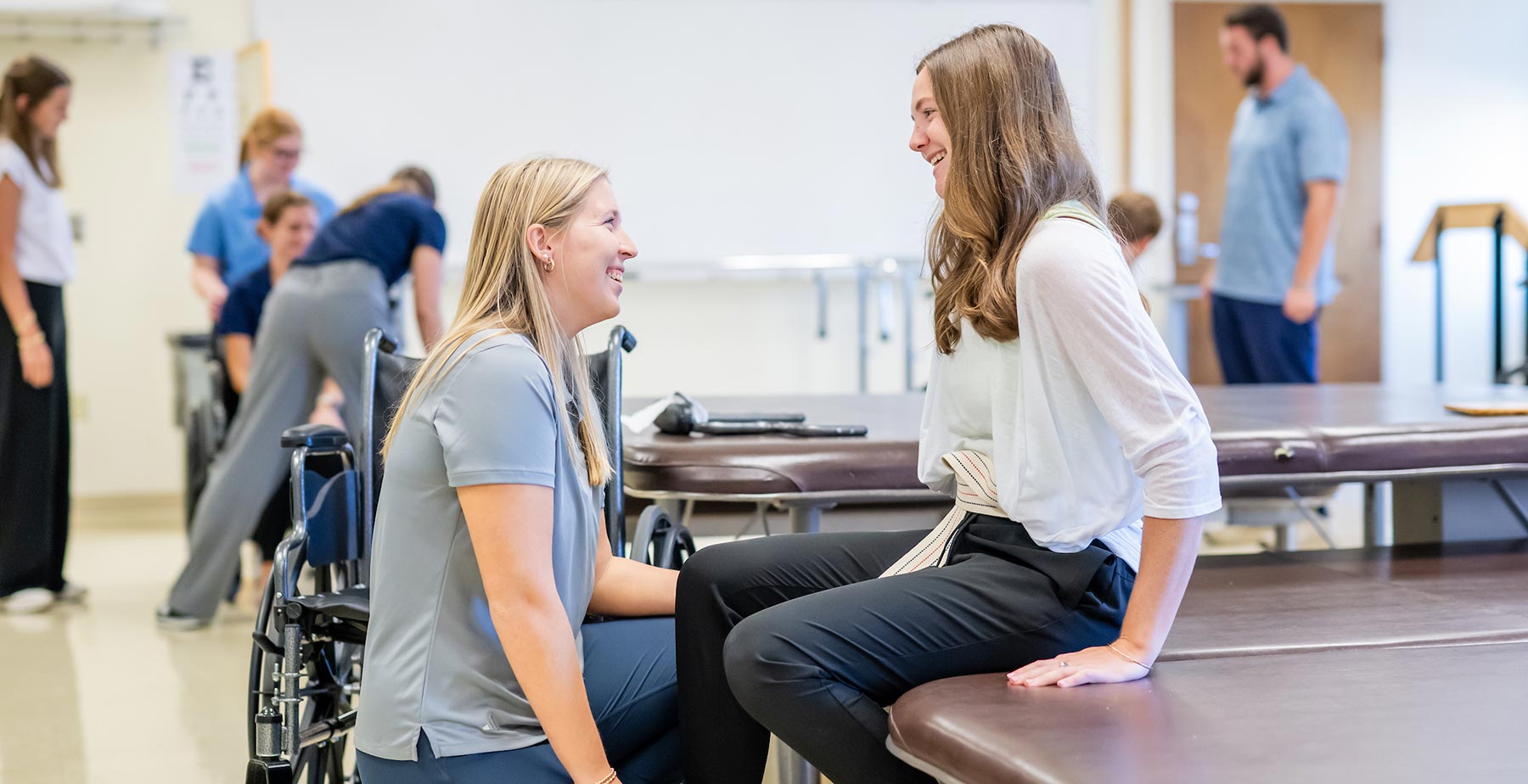A South Alabama Doctor of Physical Therapy student kneels beside a seated classmate during a hands-on clinical skills lab, with other students practicing in the background. data-lightbox='featured'