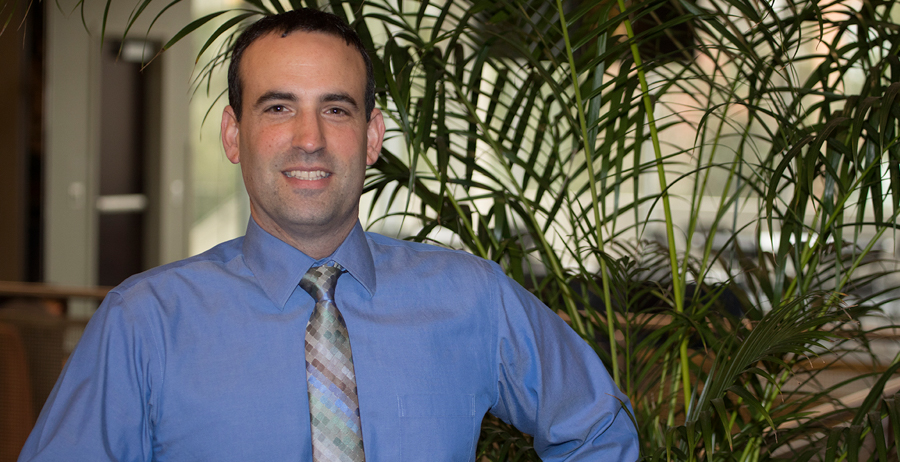 David Meola wears a blue shirt and multicolored tie, and stands in front of a backdrop of palm leaves.