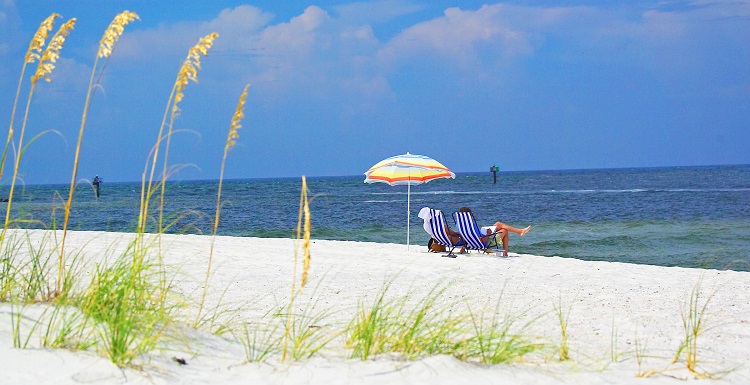 view across sand toward water of the gulf.  Sea Oats in the foreground and umbrella and beach chair in the distance