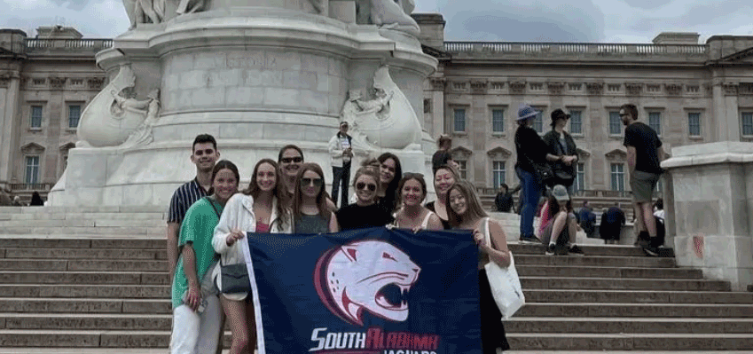 A group of BMD students stand in front of a statue of Queen Victoria in London while holding a South Alabama flag.